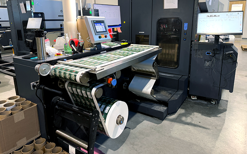 Digital HP label press printing a roll of green product labels, with control panel screens and a finished roll feeding onto the take-up reel in the production facility.