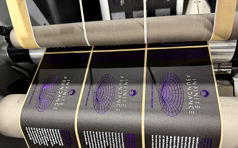 Roll of "Infinite Abundance" beer labels with a glossy black background and purple foil spiral design feeding through a label press, showing a premium finish in production.