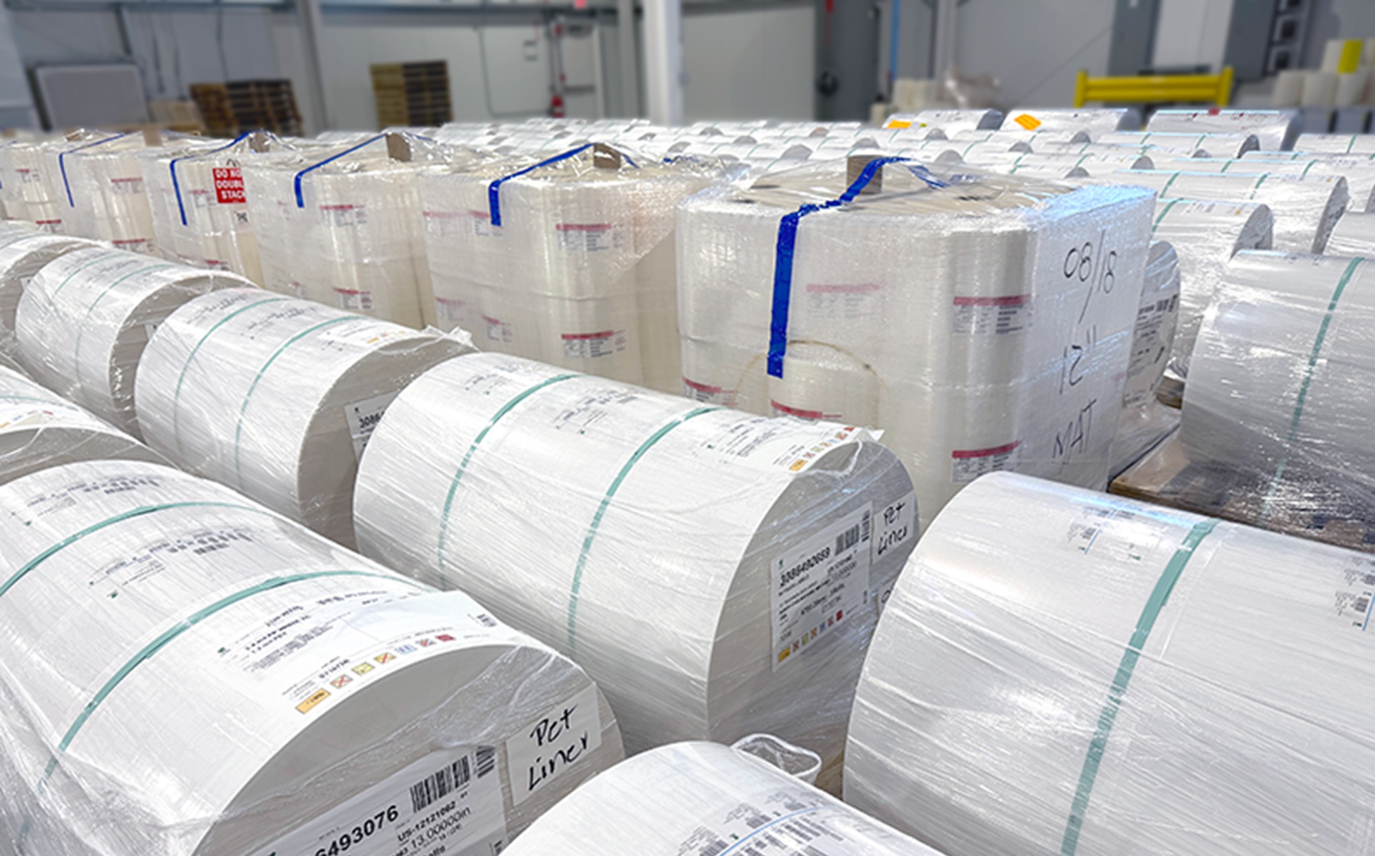 Warehouse rows of large label stock rolls wrapped in plastic, stacked and labeled with material specs like PET liner, ready for production.